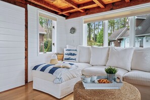 A cozy room with a wooden ceiling, white walls, a sofa, a small table with a tray of food, and a window with a view of a house and trees. - Luxury Finger Lakes Retreat - Hot Tub & Fire Pit (Cayuga)