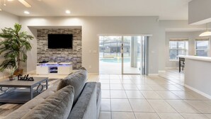 A spacious living area with a stone fireplace, a flat-screen TV, a gray sofa, and a dining area with a bar. - Modern 5BR Family Home w/ Private Pool Near Disney (Davenport)