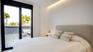 A bedroom with a large bed, two pillows, a nightstand with a lamp, and a sliding glass door leading to an outdoor patio with chairs and palm trees. - Golden Ray Sun by Interhome (Jávea)