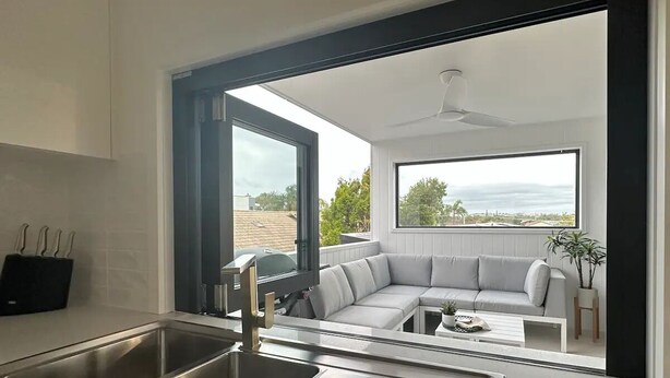 A modern kitchen with a large stainless steel sink and a view of a balcony with a gray sofa and a ceiling fan. - Coffs Harbour Beach House (Coffs Harbour)