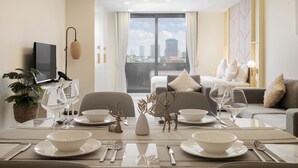 A dining area with a table set for four, including white plates, bowls, cutlery, and wine glasses. There are decorative elements like a vase and a small deer figurine. A television is mounted on the wall, and a sofa with cushions is visible in the background.