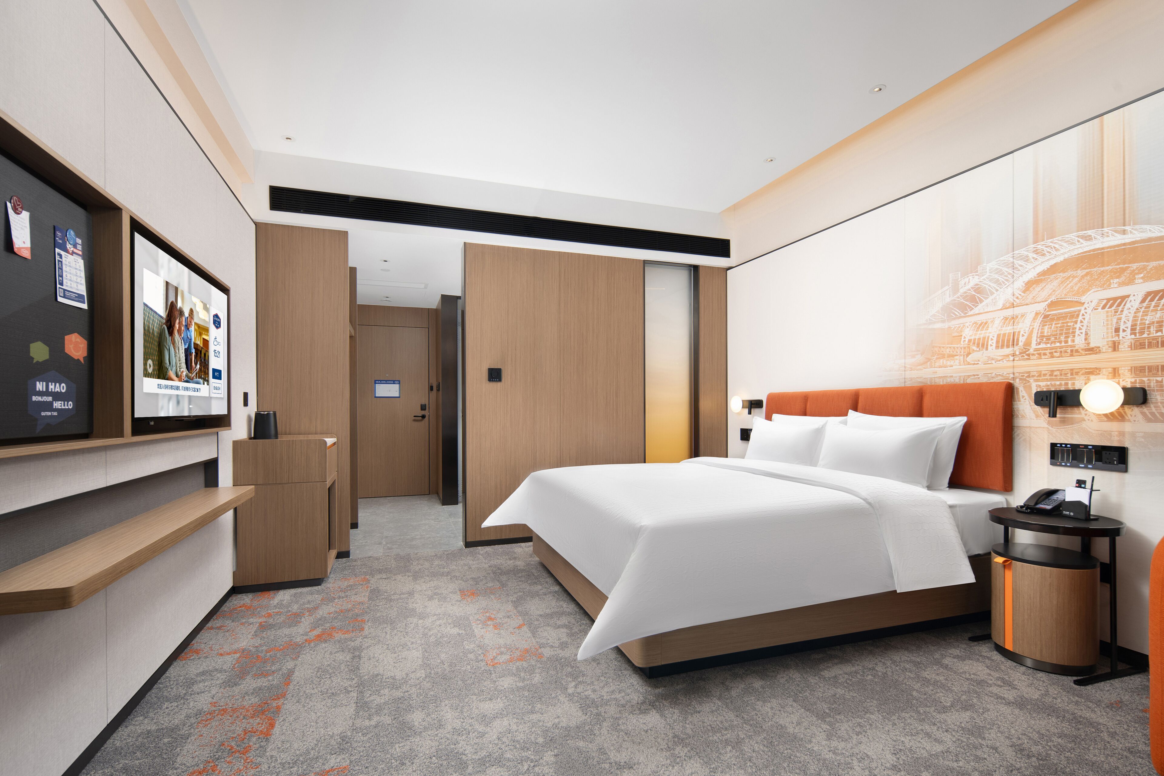 Habitación de hotel moderna con una cama grande, un escritorio y un televisor. Se observa un armario de madera y una pequeña mesa con una lámpara.