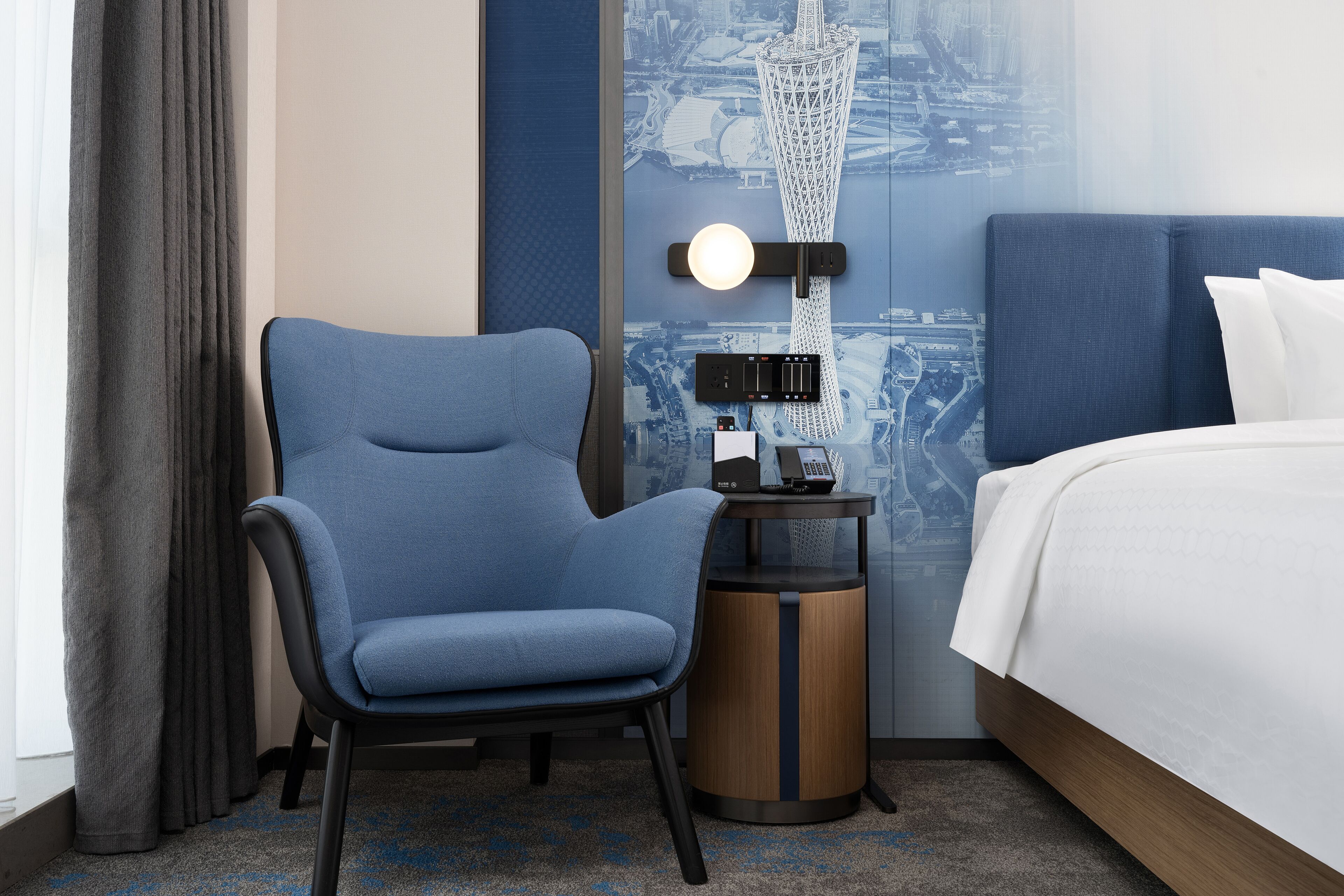 A hotel room with a blue chair, a bedside table with a lamp, and a bed with white linens.