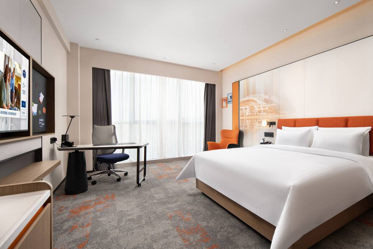 Habitación de hotel con una cama grande, un escritorio, una silla, un televisor de pantalla plana y un ventanal con cortinas.