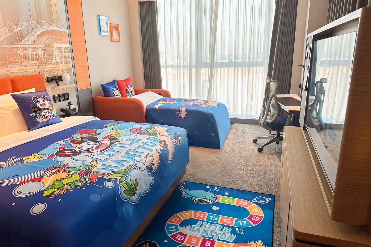 Habitación de hotel con dos camas, cada una con cubrecama de temática 'Little Hampton', un escritorio, una silla y un amplio ventanal con cortinas.