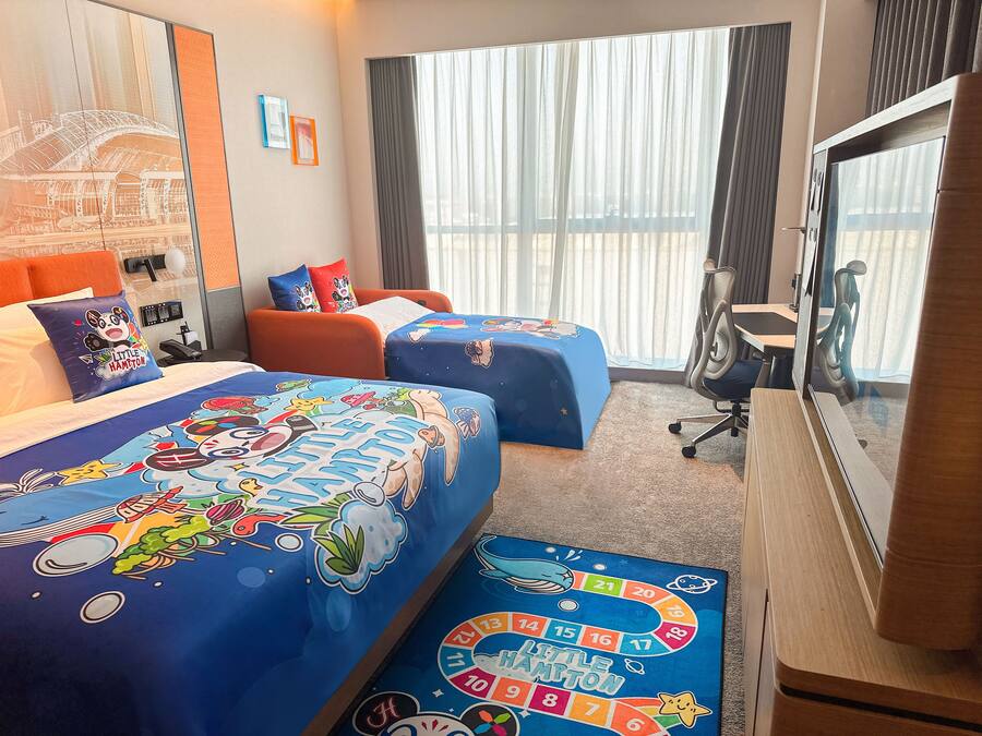 Camera d'albergo con due letti, ognuno con copriletto a tema 'Little Hampton', una scrivania, una sedia e un'ampia finestra con tende.
