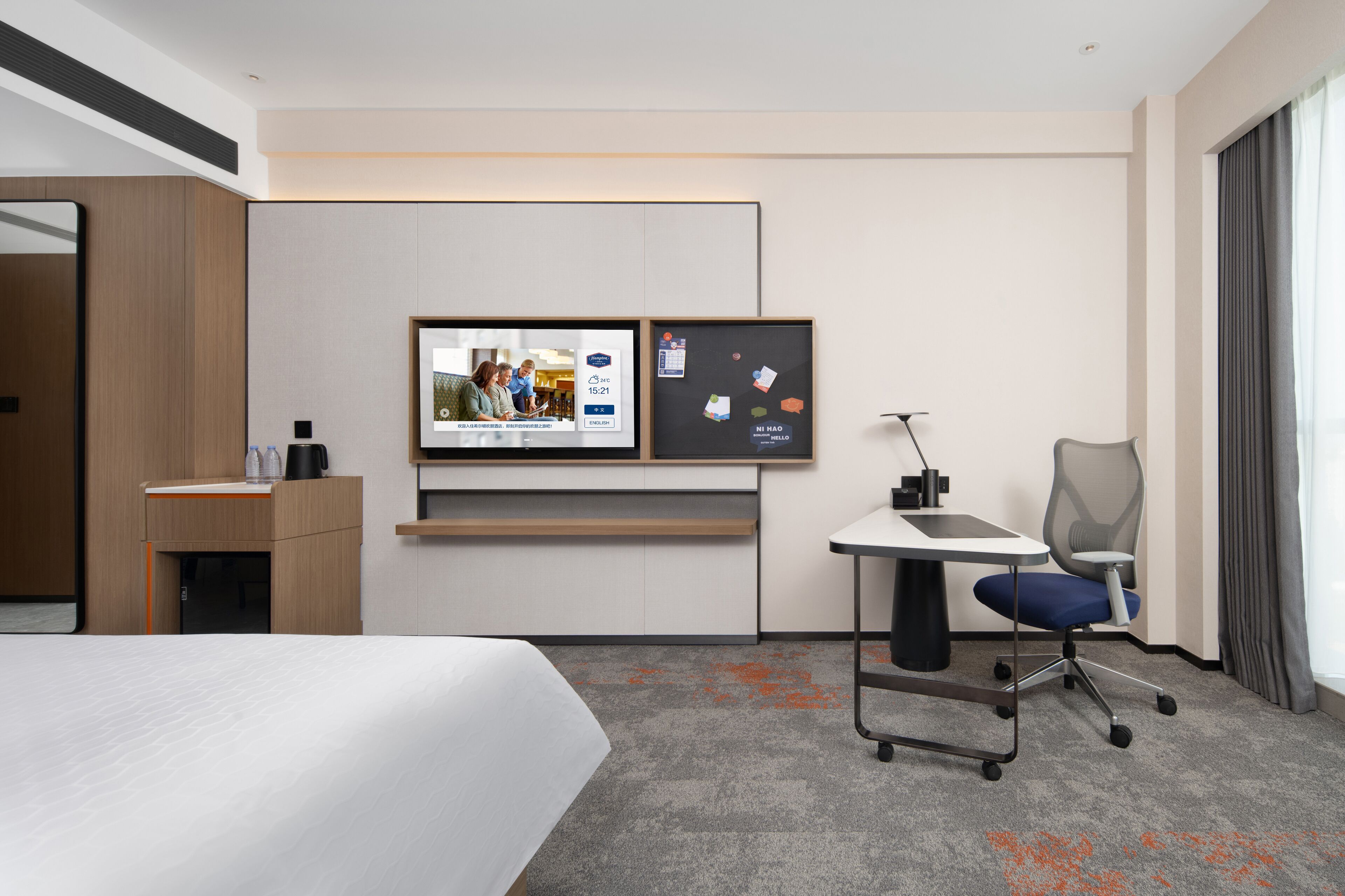 Una moderna camera d'albergo con un letto, una scrivania con una sedia, una TV fissata al muro e un'ampia finestra con tende.