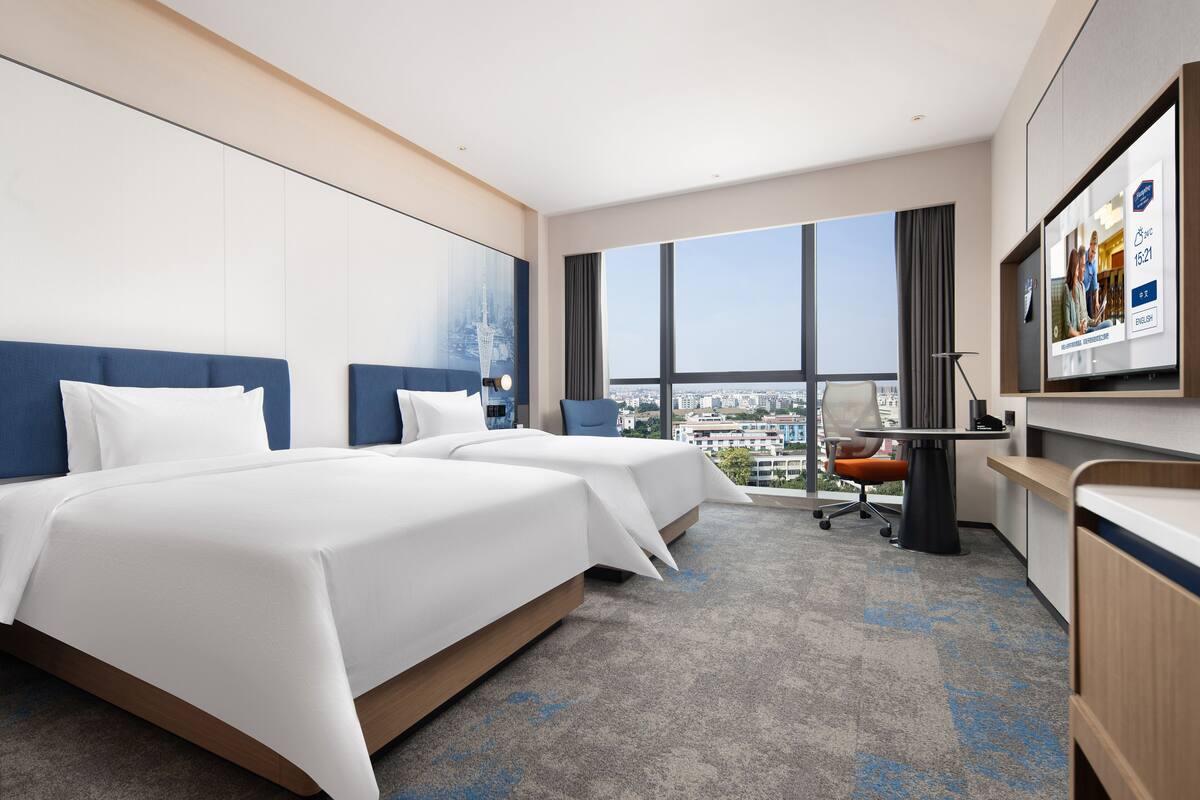 Habitación de hotel con dos camas, un escritorio, una silla y un ventanal con vistas a la ciudad.