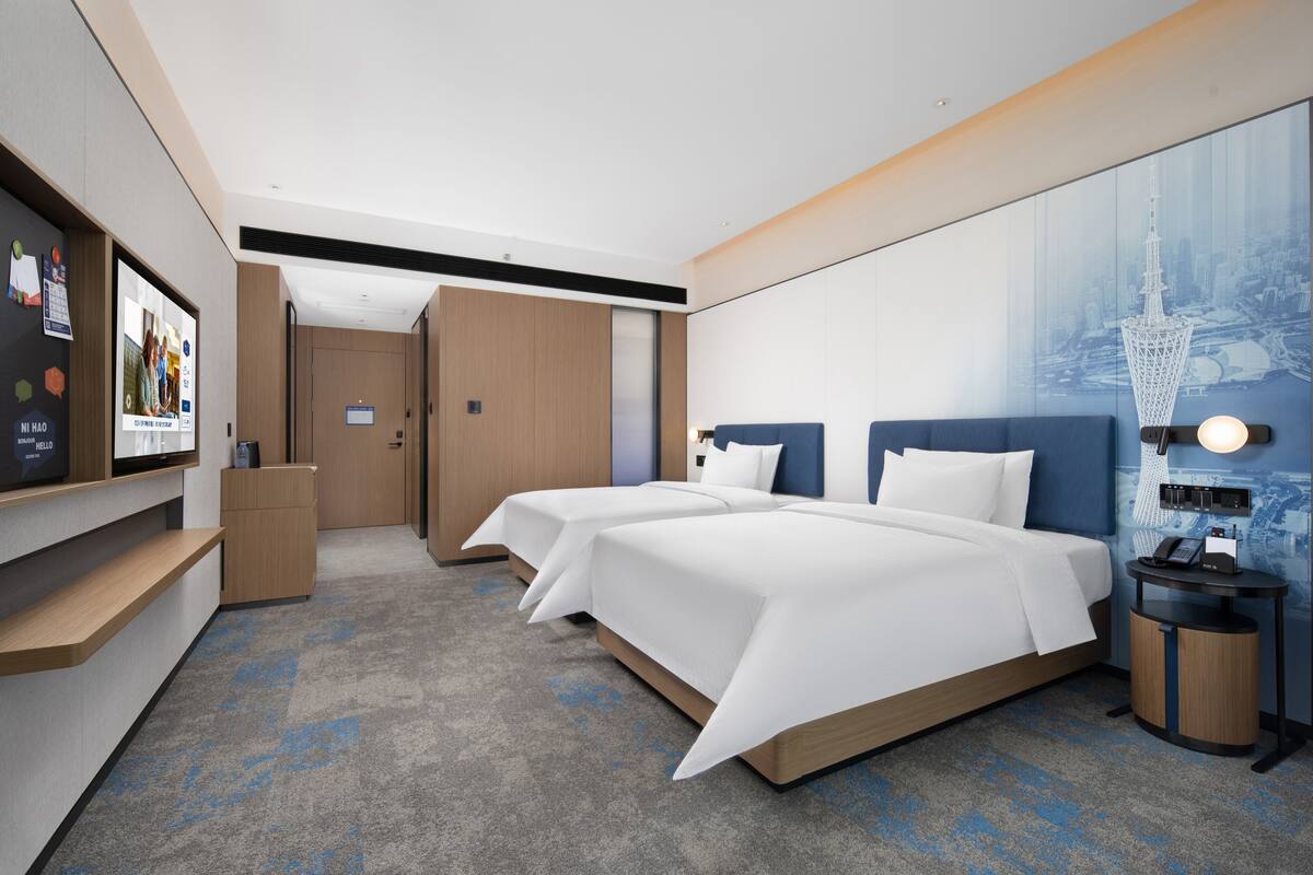 Habitación de hotel con dos camas, un escritorio, un televisor y un mural con vista a la ciudad.
