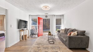 Ett modernt vardagsrum med en grå soffa, en platt-TV och en röd dörr som leder ut till en balkong.