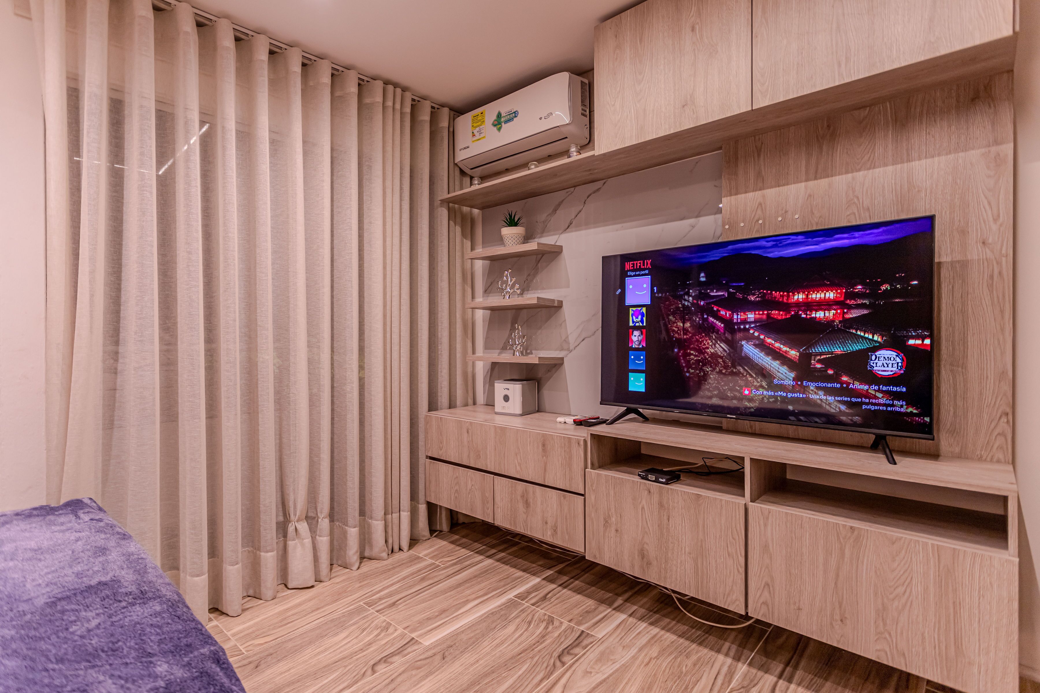 A modern living room with a flat-screen TV mounted on a wooden wall, a built-in air conditioner, and a small shelf with decorative items.