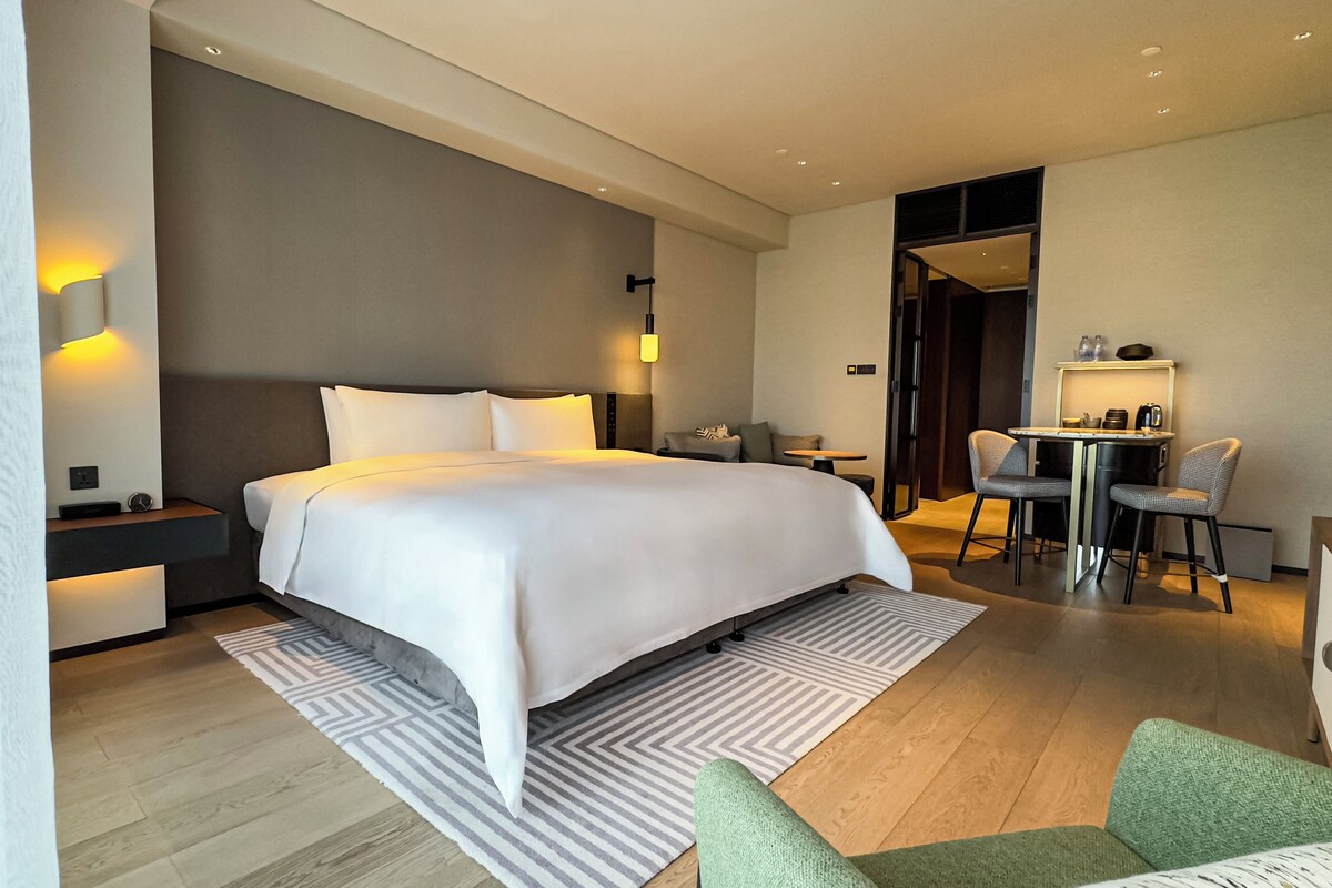 Una habitación de hotel moderna con una cama grande, mesitas de noche, un escritorio con silla y una kitchenette pequeña.