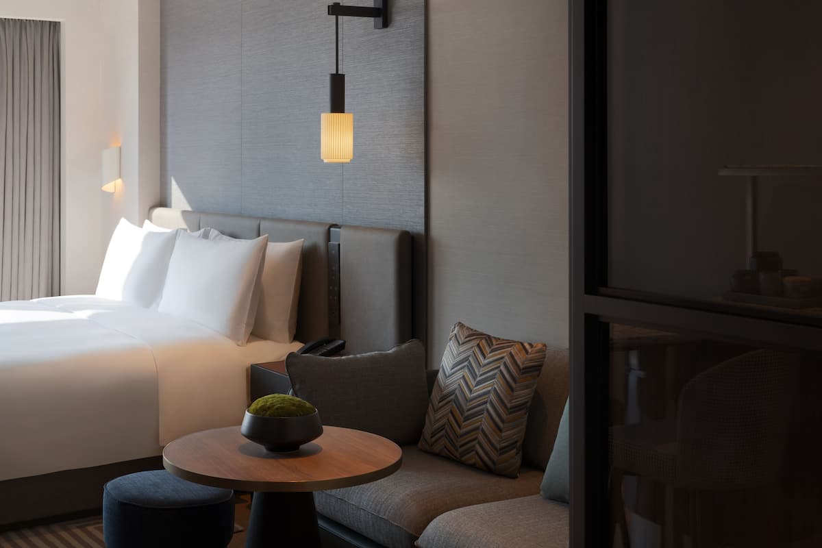 Een moderne hotelkamer met een net opgemaakt bed, een ronde houten tafel met een mosbol, een bank met decoratieve kussens en een wandlamp.