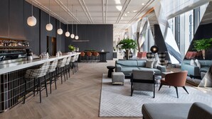 Un moderno bar con una barra larga, taburetes y una variedad de botellas. Hay una zona de estar con sofás y sillas, y ventanas amplias que ofrecen luz natural.