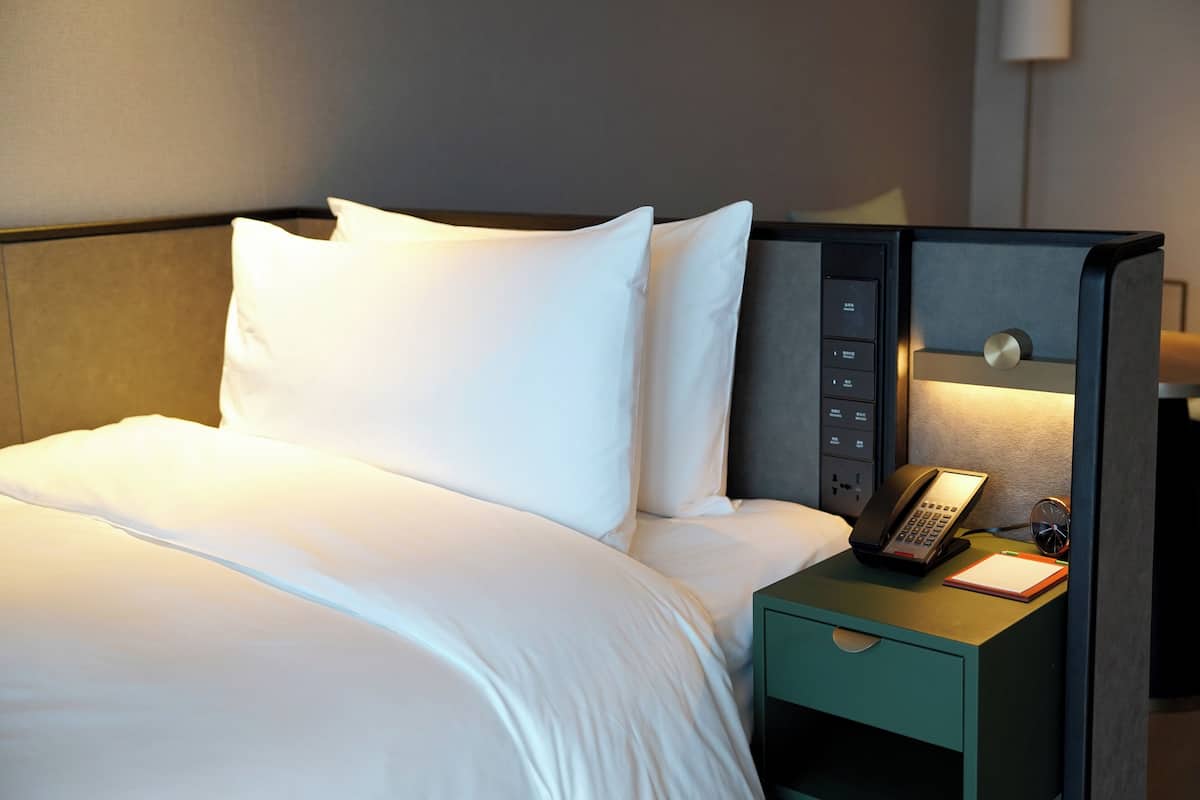 Een hotelkamer met een net opgemaakt bed, een nachtkastje met een telefoon en notitieboekje, en een wandlamp.