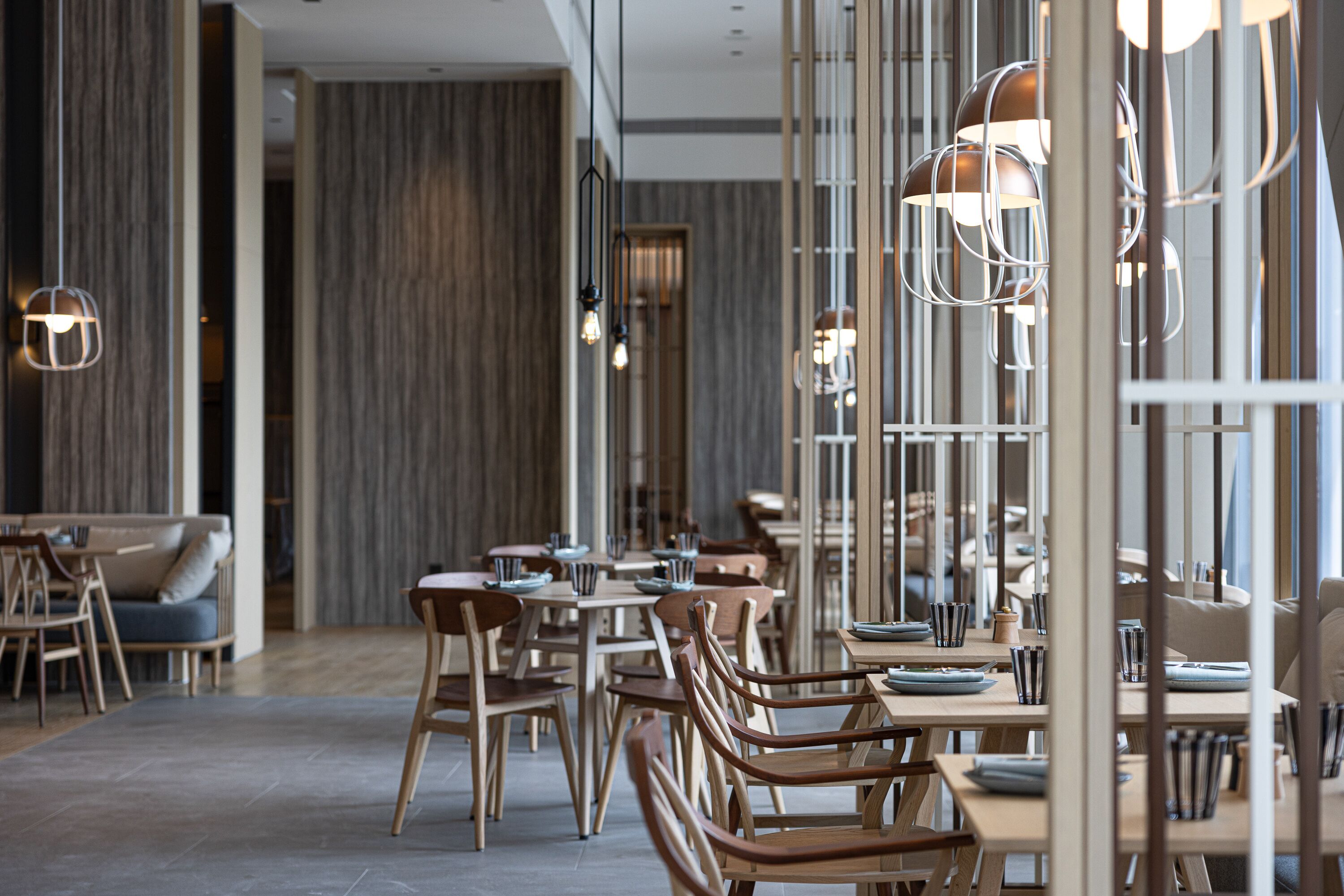 Ein modernes Restaurant mit Holztischen und -stühlen, Pendelleuchten und einer neutralen Farbgestaltung.