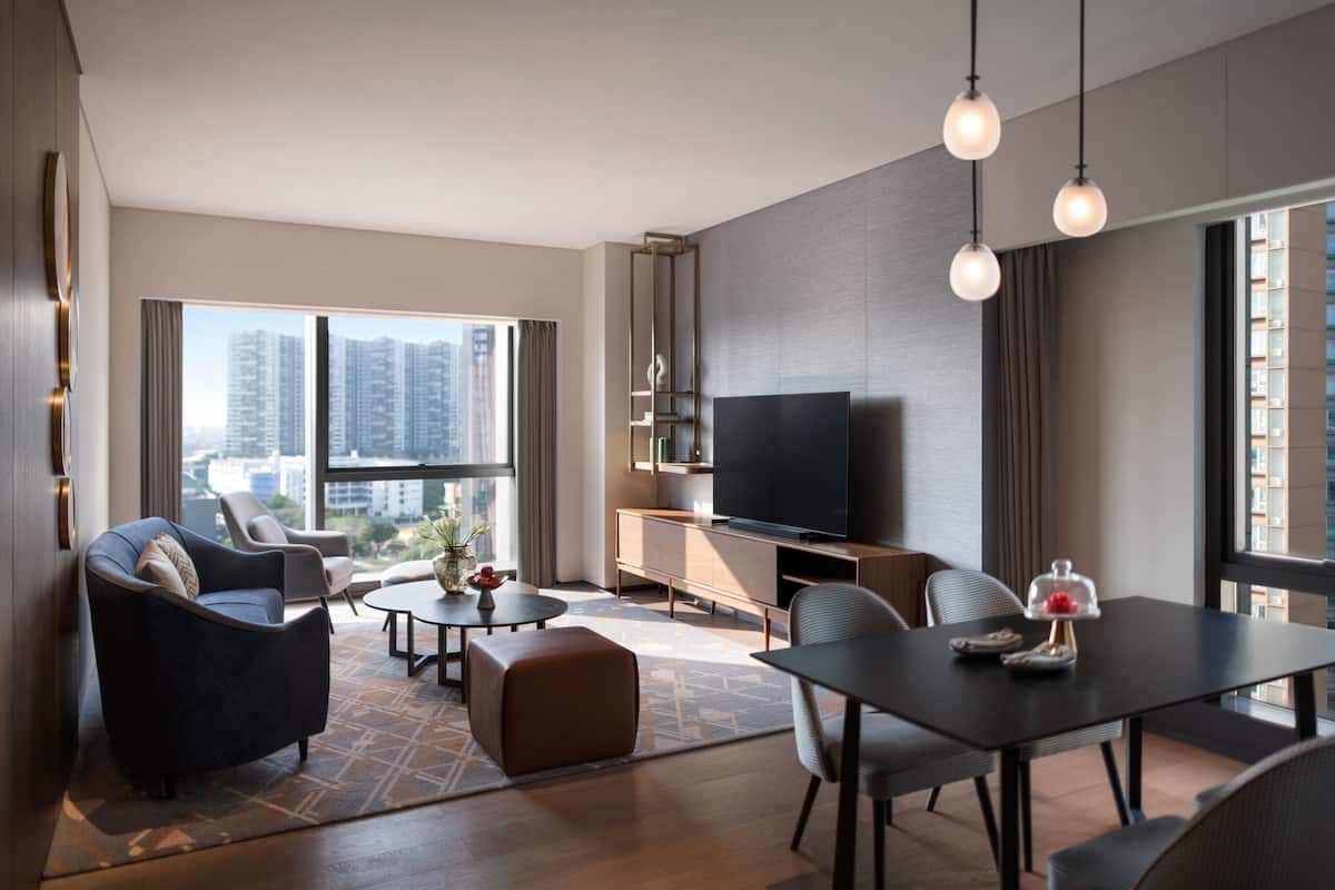 Een moderne woonkamer met een flatscreen televisie, comfortabele zitplaatsen, een eethoek en grote ramen met uitzicht op de stad.