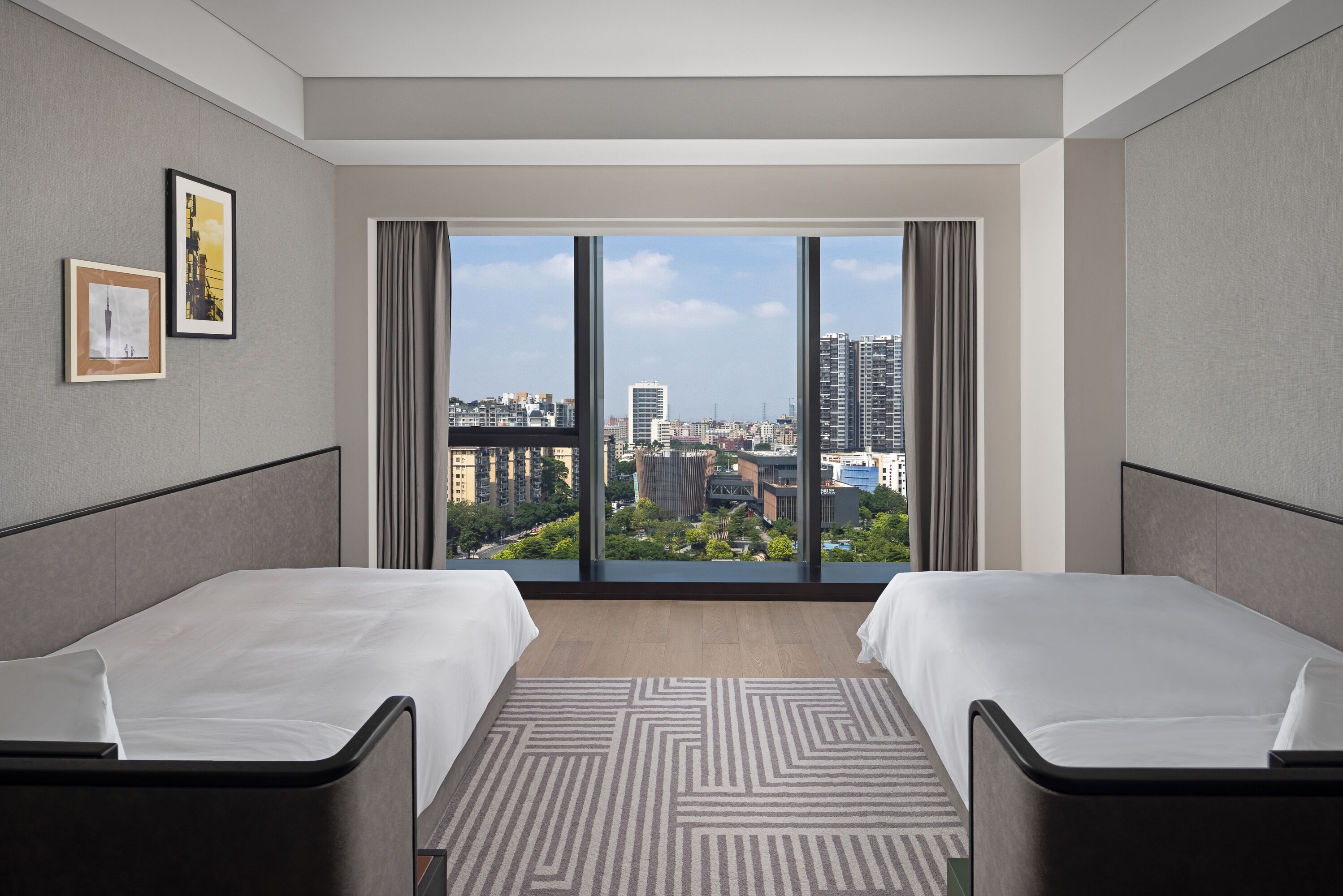 Una habitación de hotel moderna con dos camas, un ventanal con vistas a la ciudad y una alfombra estampada.