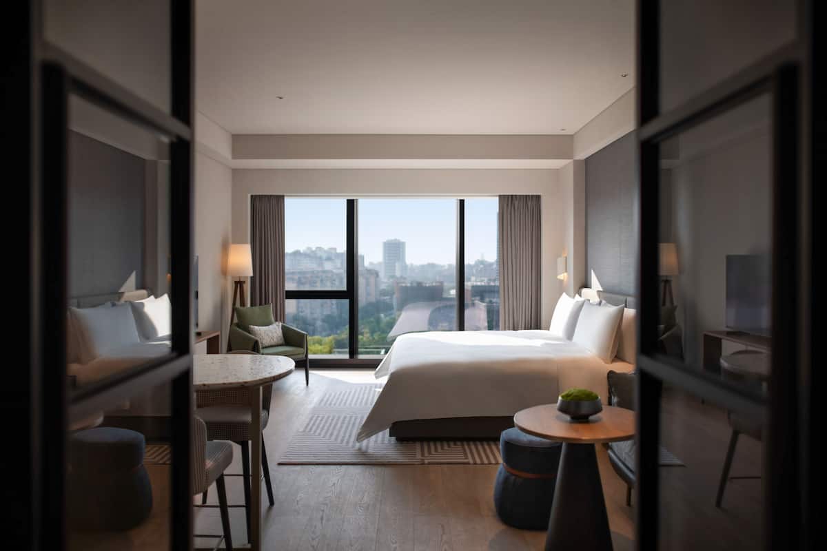 Een moderne hotelkamer met een groot bed, een zithoek met een tafel en stoelen, en een stadszicht door grote ramen.