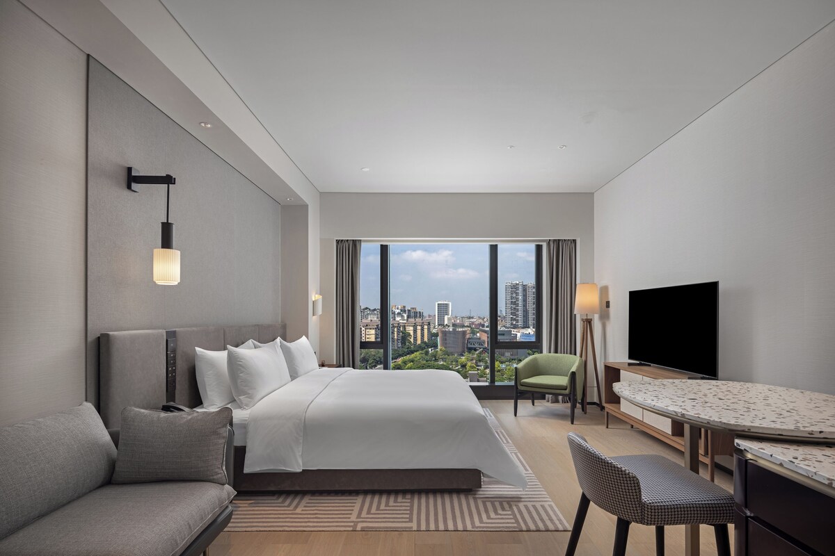 A modern hotel room with a large bed, a sofa, a dining table, and a TV.