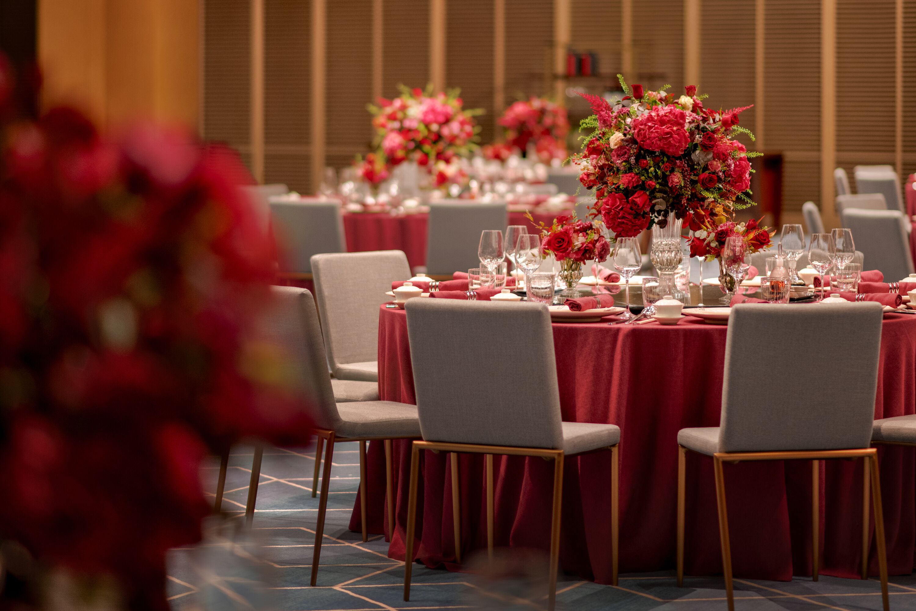 Une salle de banquet avec des tables rondes dressées pour le repas, chacune étant recouverte d’une nappe rouge et agrémentée de compositions florales.