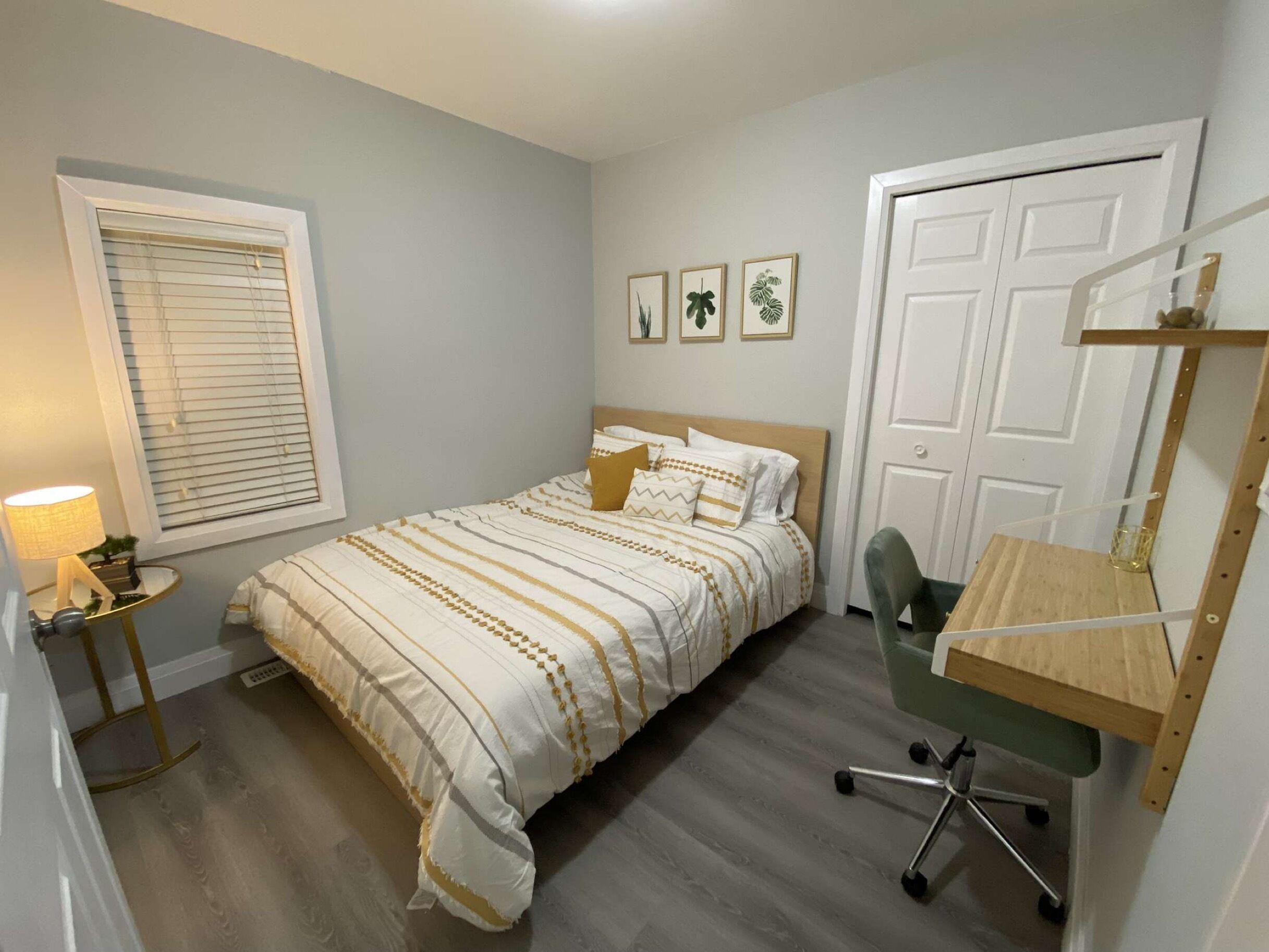 A bedroom with a bed, a desk, a chair, a window with blinds, and a door.