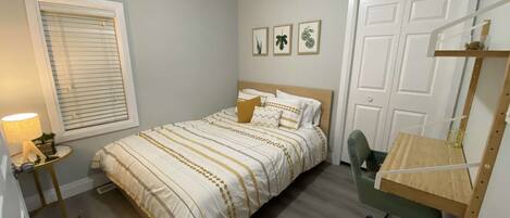 A bedroom with a bed, a desk, a chair, a window with blinds, and a door.