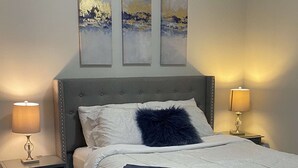 Un llit ben fet amb un capçal gris, roba de llit blanca i una manta blava. Hi ha dues taules de nit amb làmpades i tres quadres abstractes penjats a la pared sobre el llit.