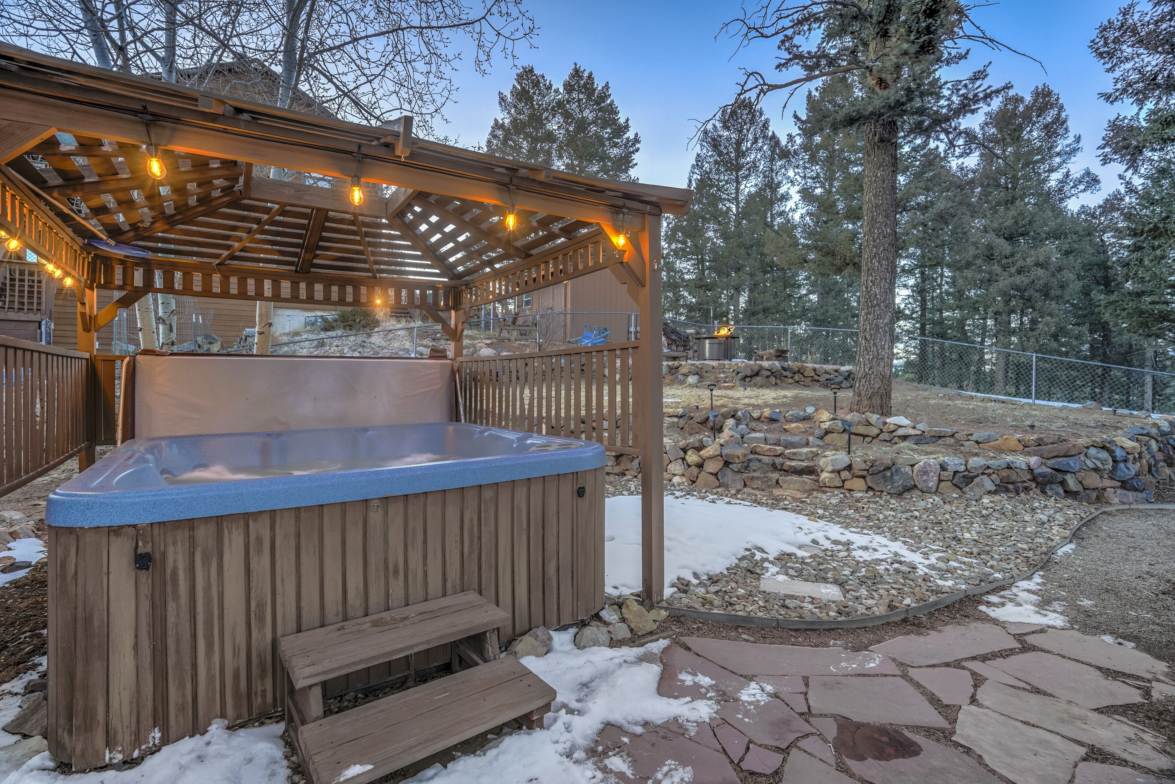 A hot tub under a wooden pergola with string lights, surrounded by a stone wall and a wooden deck.