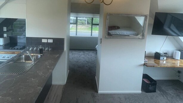 A compact kitchen with a built-in stove, a sink, and a chandelier. There is a small television on a wooden shelf and a window providing natural light. - Ohope Dreamer 1 (Ōhope)