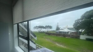 A room with a large window overlooking a grassy yard, a metal staircase, and a white building in the background. - Ohope Dreamer 1 (Ōhope)