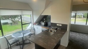 A compact kitchen with a central island, a dining area with a glass table, and a view of the outdoors. - Ohope Dreamer 1 (Ōhope)