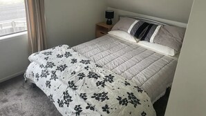 A bedroom with a bed, a nightstand, a lamp, and a window with curtains. - Ohope Dreamer 1 (Ōhope)