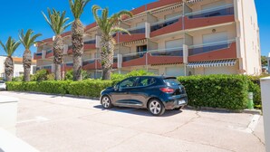 Un edificio residencial de varios pisos con balcones, palmeras y un automóvil estacionado.