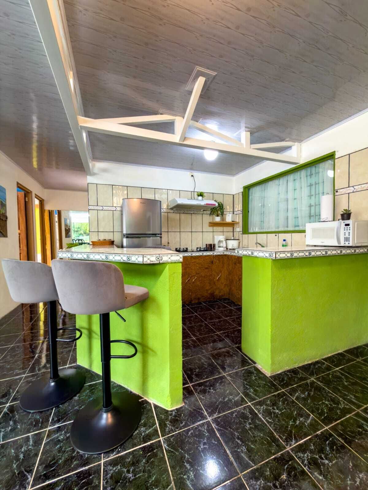 A kitchen with a green island, two bar stools, a stainless steel refrigerator, a microwave, and a window with curtains.