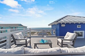 A deck with two white cushioned chairs and a small table, overlooking a beach and a blue building. - North Star | Ocean Views + Rooftop! (Carolina Beach)