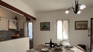 A dining area with a wooden table set for four, surrounded by white chairs. A chandelier hangs above, and there are two windows with curtains. - Casa Iose by Interhome (Torrazza)