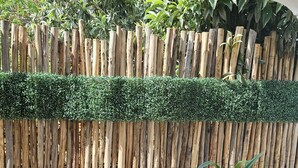 Una cerca de madera con parte superior verde, una jardinera con plantas y un suelo de baldosas.