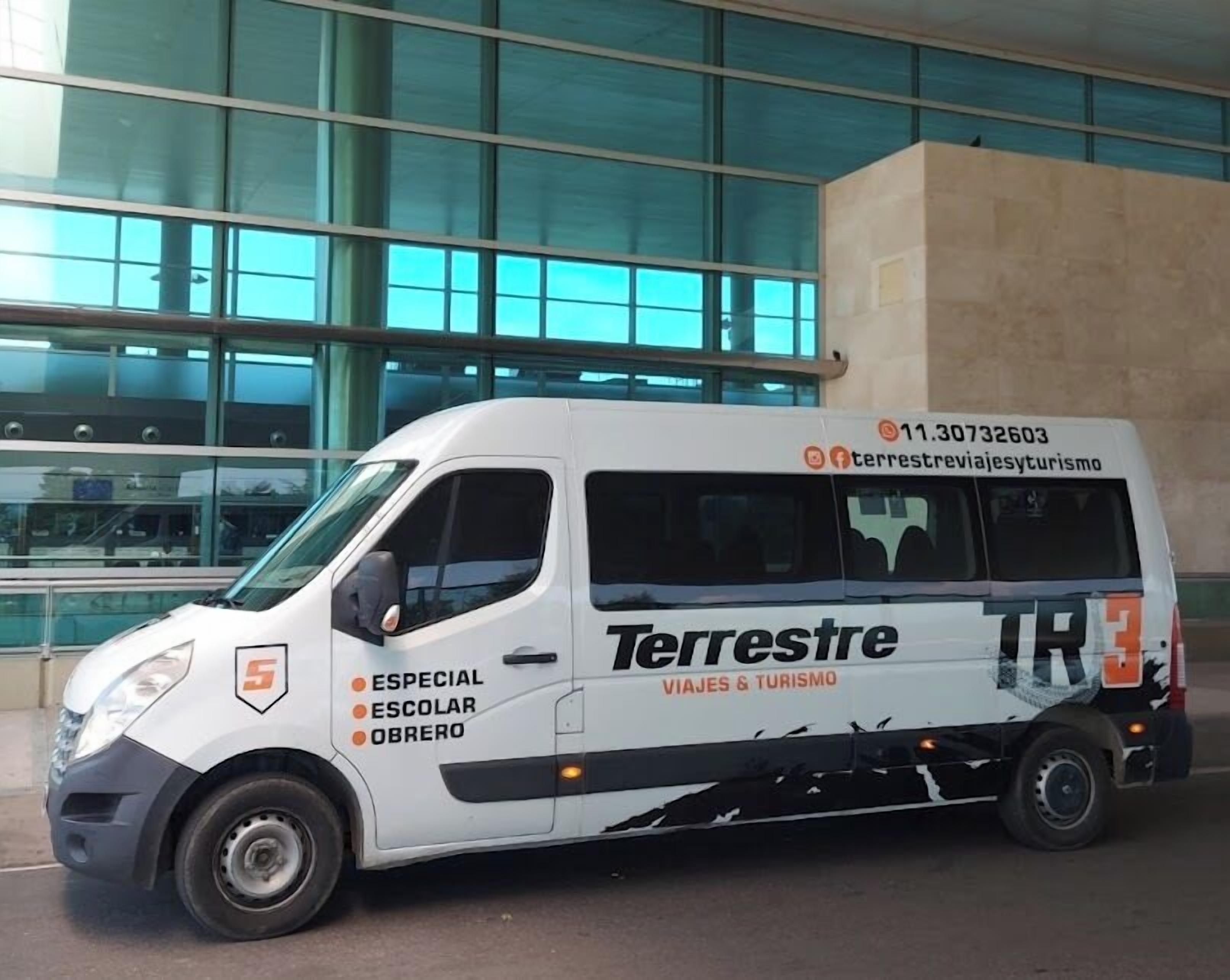 A white van with 'Terrestre' branding and 'VIAJES & TURISMO' text, parked in front of a modern building with large windows.