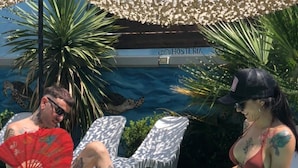 Dos personas sentadas junto a una piscina, con sillones de descanso y un letrero que dice "HOSTELIA" al fondo.
