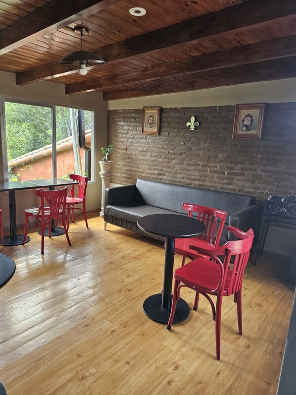 Una habitación acogedora con techo de madera, pared de ladrillo, sofá gris, mesa de comedor con sillas rojas y una planta en maceta.