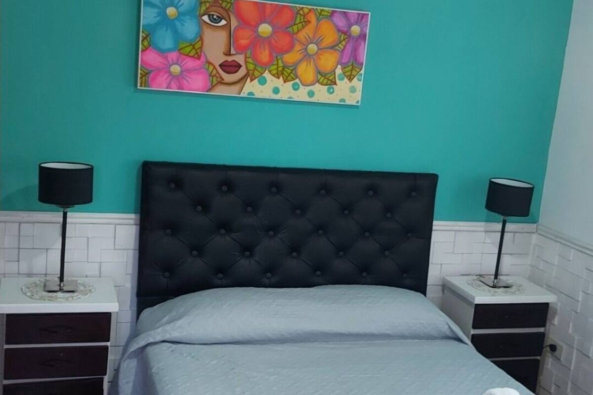 Un dormitorio con pared color turquesa, cabecera tapizada negra, cama con colcha blanca, dos mesitas de noche con lámparas y un cuadro colorido sobre la cama.