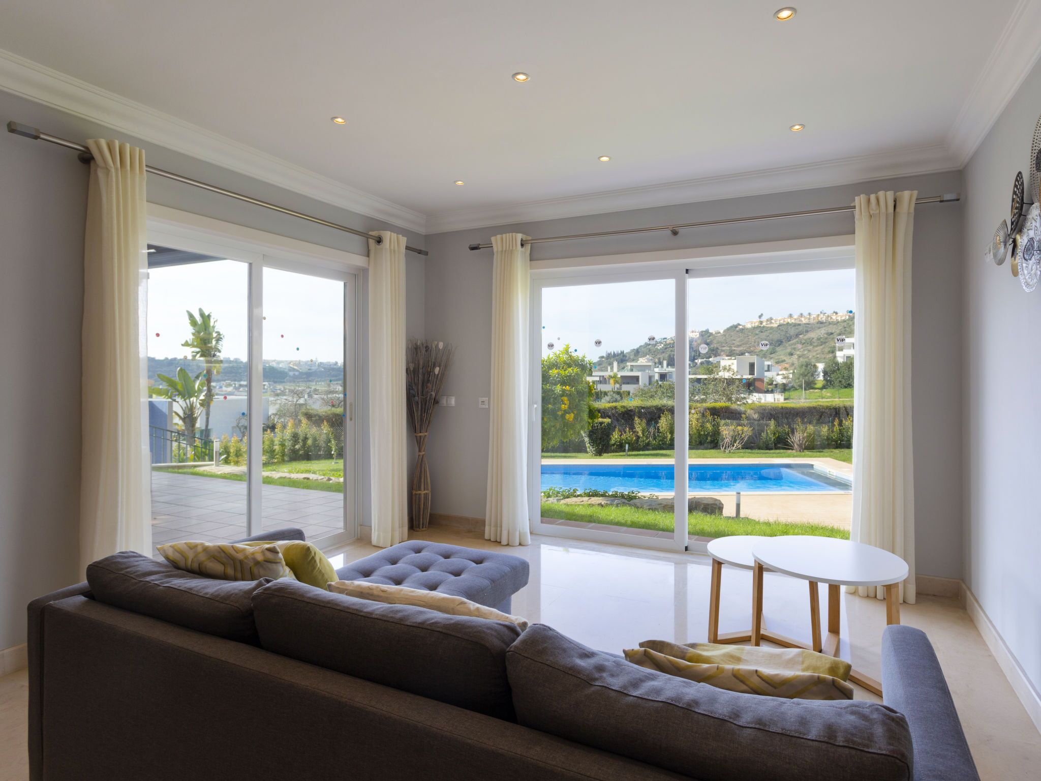 A spacious living room with a large sofa, a small round table, and a view of a swimming pool and greenery.