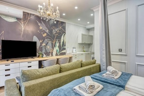 A modern hotel room with a flat-screen TV, a sofa, a bed with towels, a chandelier, and a kitchen area. - Boutique Downtown Studio with Balcony 1 (Zagreb)
