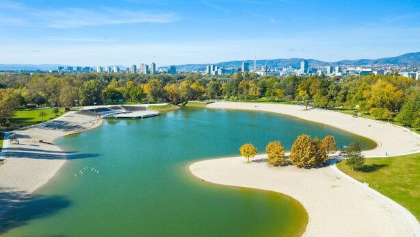 An aerial view of a park with a lake, sandy areas, and a cityscape in the background.