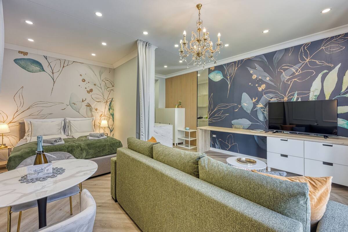 Un salon moderne avec un canapé vert, une table basse blanche, un lit avec une literie verte, un lustre et un téléviseur.