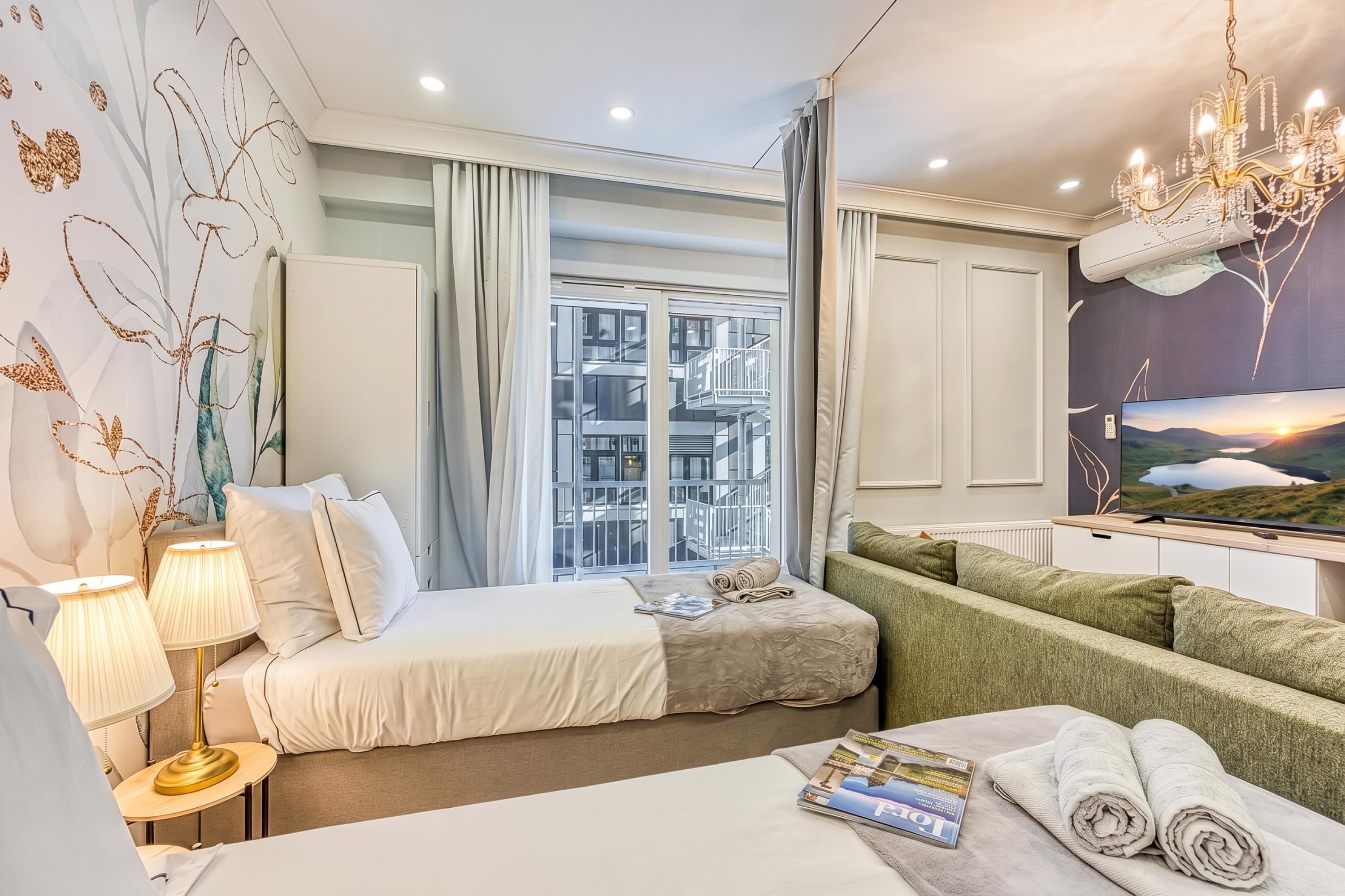 A hotel room with a bed, a sofa, a desk with a lamp, a TV, and a window with a view of buildings.