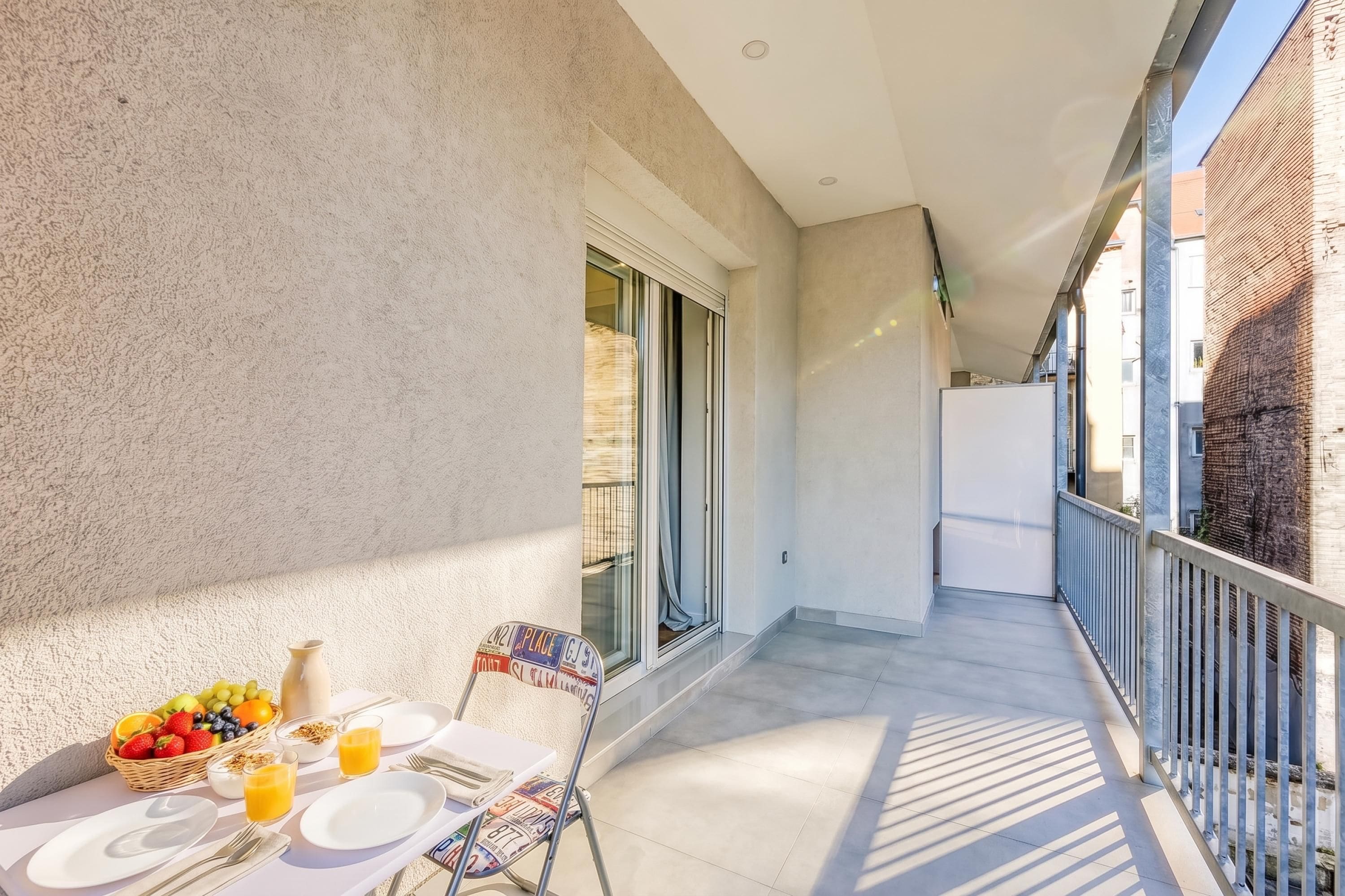 A balcony with a table set for breakfast, including fruits, juice, and a vase.