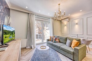 A modern living room with a grey sofa, a flat-screen TV, a chandelier, and a large window with curtains. - Boutique Downtown Studio with Balcony 1 (Zagreb)