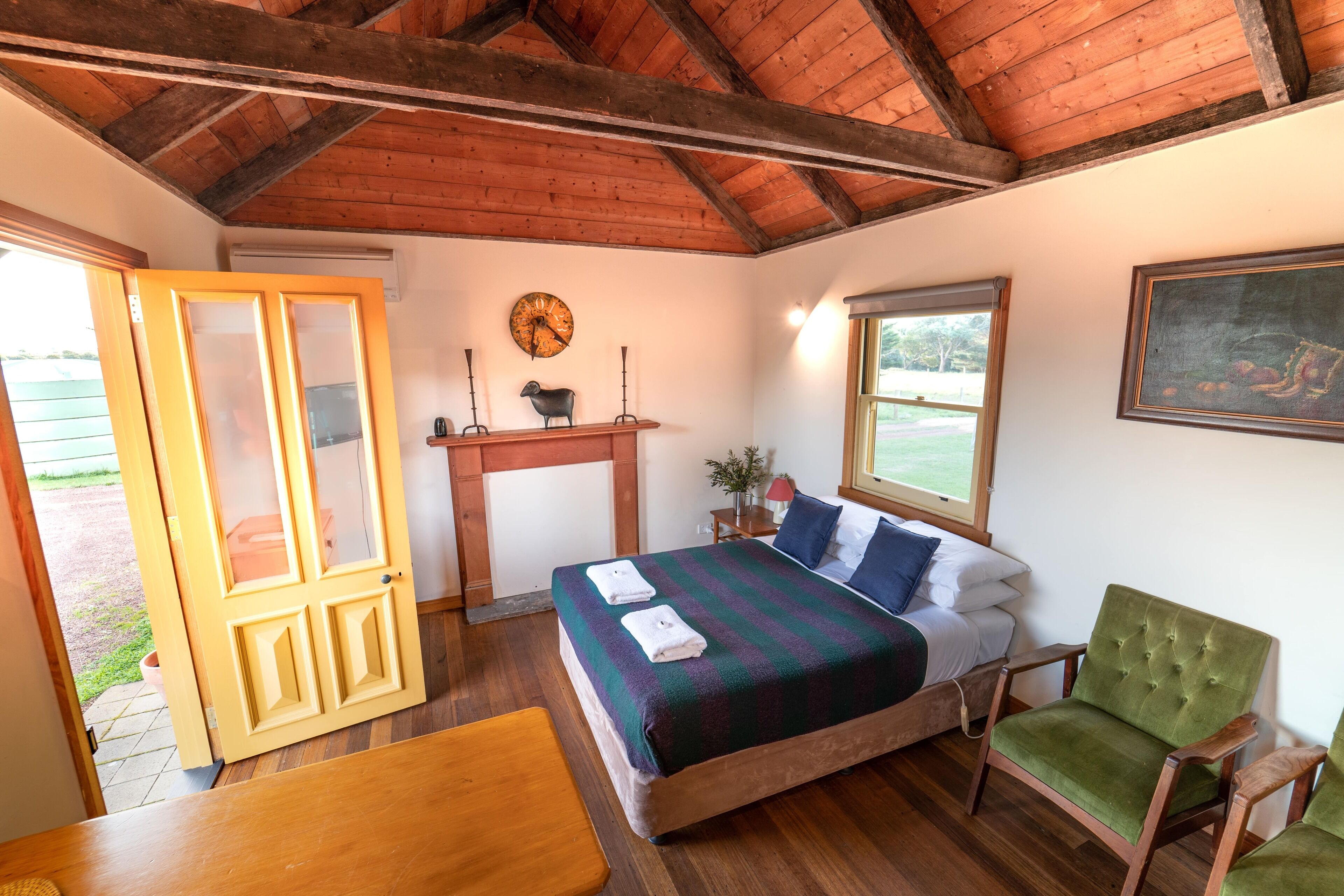 A bedroom with a wooden ceiling, a bed with a striped blanket, a green armchair, a wooden nightstand, and a window with a view of the outdoors.